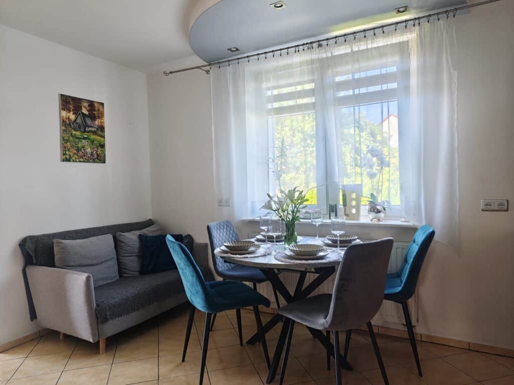 Kącik jadalny w apartamencie Sweet Stay z nakrytym stołem, niebieskimi krzesłami, szarą sofą i dużym oknem z firanami